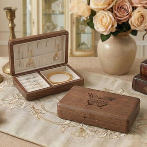 May include: Open wooden jewelry box with a light-colored interior, displaying necklaces, rings, and a bracelet. A closed wooden box with a butterfly design and the name "Sophia" is next to it. The boxes are on a patterned surface.