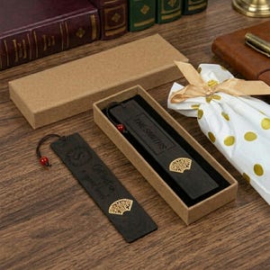 May include: Two dark wooden bookmarks with gold fan designs and red beads. One bookmark is in a cardboard box, and the other is outside the box. One bookmark has the text "THE SMITHS" and the other has the text "Get lost in a good book".