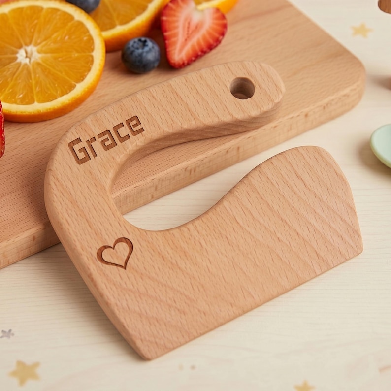 Close up of a custom kids kitchen tool set showing an engraved junior chef slicer personalized with the name Grace and a cute heart icon. The wooden toddler knife rests on a matching cutting board surrounded by fresh fruit in a warm, cozy setting. This Waldorf fine motor skill toy highlights natural wood grain and precise laser engraving.