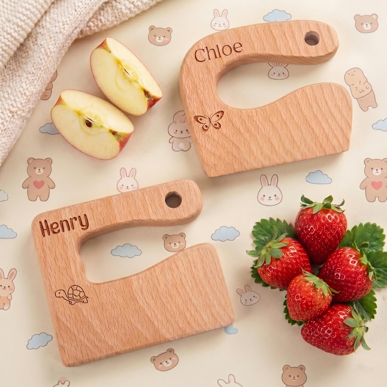 Two custom engraved junior chef slicers displayed on a cute animal background with fresh strawberries and apples. One wooden knife is personalized for Chloe with a butterfly, and the other for Henry with a turtle. Great Waldorf fine motor skill toy options.
