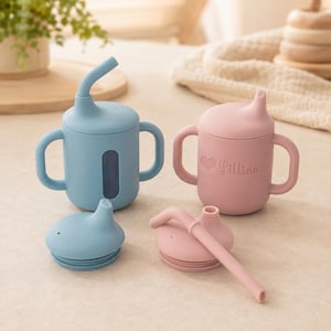 Könnte beinhalten: Zwei Silikon-Trinklernbecher mit Griffen, einer in Blau und einer in Rosa. Der blaue Becher hat einen Strohhalmdeckel und ein vertikales Sichtfenster. Der rosa Becher zeigt ein Herzsymbol und den Namen Lillian. Zwei Ersatzdeckel liegen bei.