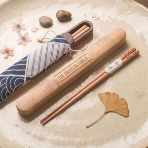 May include: A pair of light-colored wooden chopsticks with engraved text reading THE ANDERSO and EST. 2026, accompanied by a matching wooden storage case engraved with THE ANDERSON FAMILY and a blue and white patterned fabric pouch.