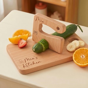 Custom kids kitchen tool set in action. A personalized wooden chopper engraved with Mia and a bow icon slices through a cucumber on a matching Mia's Kitchen cutting board. Surrounded by fresh fruit, this Montessori safe slicer makes a perfect toddler gift.