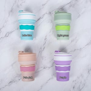 A clean, top-down flat lay view displaying the four color options for the personalized collapsible silicone water cups on a marble surface. The set includes Lake Blue, Light Green, Pale Pink, and Purple, each featuring a silicone bottle band.