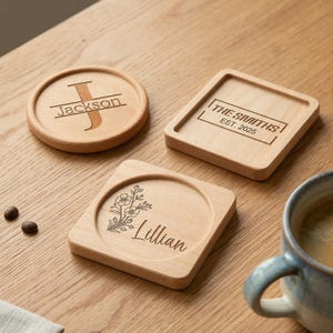 May include: Wooden coasters with engraved designs. One round coaster features the name "Jackson" with a letter "J". A square coaster has "The Smiths EST. 2025". Another square coaster has the name "Lillian" with a floral design. A blue mug is on the right.