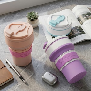 A lifestyle photo of personalized collapsible silicone water cups on a marble desk. Two fully extended cups feature engraved name bands, "Chloe" in pink and "Lillian" in purple. Other collapsed and folded cup is present, showing the range of sizes. Also shown are a notebook, pen, a succulent plant, and wireless earbuds case, suggesting portable use.