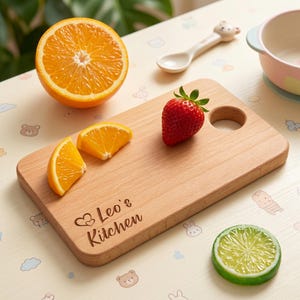 A personalized wooden cutting board from the custom kids kitchen tool set resting on a cute patterned mat. Engraved with Leo's Kitchen and a double heart icon. Displayed alongside fresh oranges and a strawberry, highlighting this toddler chef accessory.