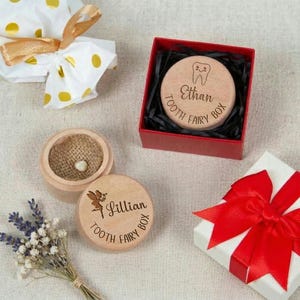 Op de afbeelding: Houten tandfee-dozen met gegraveerde namen en ontwerpen. Eén doos is open en onthult een tand. Een andere doos zit in een rode doos met zwarte vulling. Een klein boeket lavendel en een cadeau met een rode strik zijn ook aanwezig.