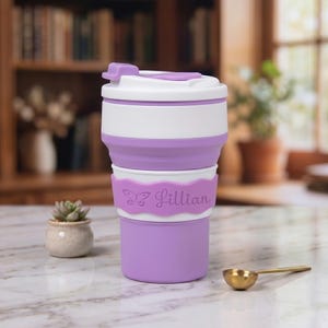 A close-up portrait of the personalized purple collapsible silicone water cup on a kitchen marble countertop. The neon purple name band is elegantly engraved with a butterfly icon and the name "Lillian" in a delicate script. Ideal for coffee or a unique gift.