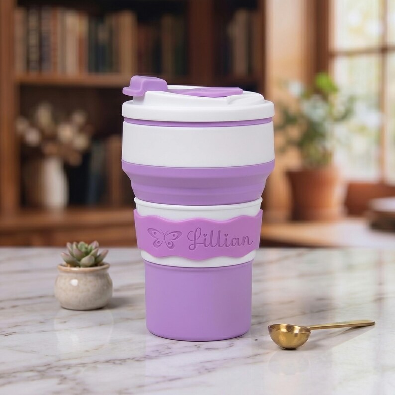 A close-up portrait of the personalized purple collapsible silicone water cup on a kitchen marble countertop. The neon purple name band is elegantly engraved with a butterfly icon and the name "Lillian" in a delicate script. Ideal for coffee or a unique gift.