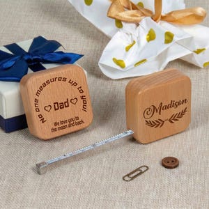 Può includere: Due metri a nastro in legno con testo inciso. Uno recita "Nessuno ti misura! Papà Ti amiamo fino alla luna e ritorno". L'altro dice "Madison" con un disegno di foglie. Un nastro di metallo si estende da uno.