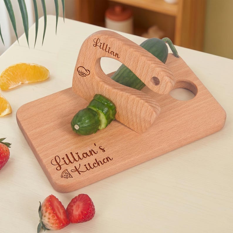 Close-up view of a custom engraved junior chef slicer safely cutting a green cucumber on a wooden board. The toddler-safe wooden knife is personalized with Lillian and a chef hat. An ideal Waldorf fine motor skill toy for little chefs prepping food.