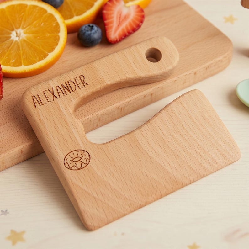 Detailed view of a personalized wooden knife from our custom kids kitchen tool set. This engraved junior chef slicer features the name Alexander in bold letters with a fun donut icon. Set against a warm, professional backdrop with fresh fruit, this beautiful Waldorf fine motor skill toy showcases realistic wood textures and custom details.