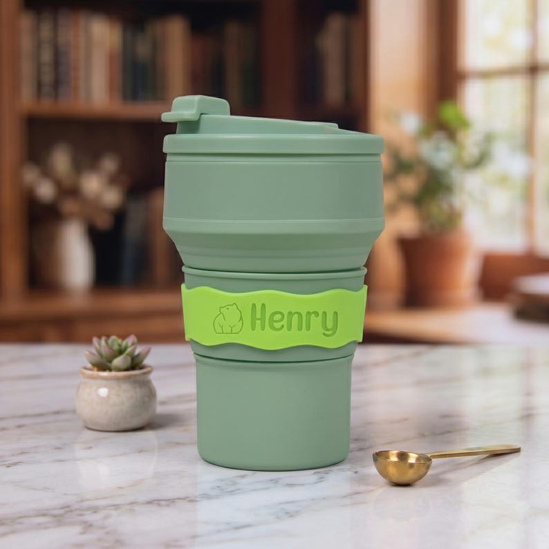 A close-up view of the light green personalized collapsible silicone water cup on a marble table. The bright neon green name band features a custom engraved bear icon and the name "Henry" in a modern font. It highlights the vibrant color options for kids or outdoor enthusiasts.