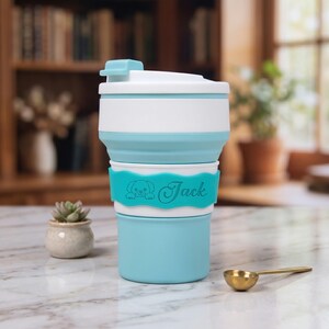 A close-up, full view of the personalized lake blue collapsible silicone water cup on a kitchen marble countertop. The name band clearly shows the engraved dog icon and the name "Jack." A small succulent and a gold scoop are nearby, all with a blurred background of a bookshelf and plants, creating a cozy and practical setting.