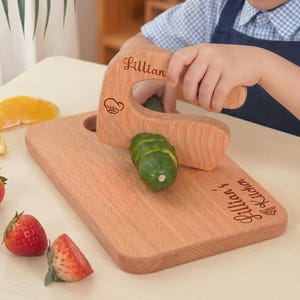 A young child wearing an apron uses an engraved junior chef slicer to cut a cucumber. The custom wooden knife and cutting board are personalized with the name Lillian. This Waldorf fine motor skill toy is a safe way for toddlers to learn cooking.