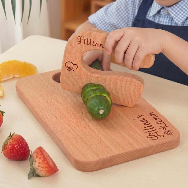 A young child wearing an apron uses an engraved junior chef slicer to cut a cucumber. The custom wooden knife and cutting board are personalized with the name Lillian. This Waldorf fine motor skill toy is a safe way for toddlers to learn cooking.