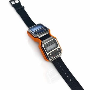 Pode incluir: Um relógio digital de estilo retrô com uma pulseira de tecido preto e uma caixa laranja e prateada. O mostrador do relógio exibe a data e a hora. O relógio tem formato retangular com bordas arredondadas.