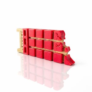 Puede incluir: Primer plano de un objeto rojo y beige con un patrón de rejilla. El objeto tiene una serie de bloques cuadrados rojos dispuestos en filas, sobre un fondo beige. El objeto está sobre una superficie blanca.