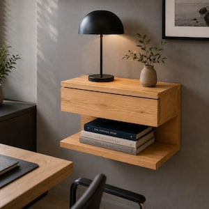 May include: A wall-mounted light oak floating nightstand featuring a single drawer and an open lower shelf. The unit holds a black dome-shaped table lamp, a small ceramic vase with green foliage, and three stacked books with visible text.