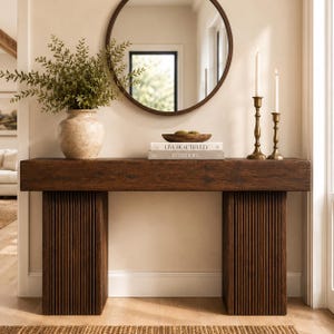 Op de afbeelding: Een donkerbruine houten sidetable met een dik rechthoekig blad en twee gecanneleerde zuilvormige poten. Op de tafel staan een beige keramische vaas met groen blad, een houten schaal, twee gestapelde boeken met de titels Live Beautifully en Interiors, en twee koperen kandelaars.