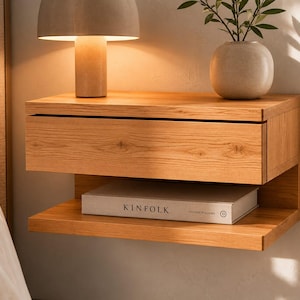 May include: A wall-mounted light oak nightstand featuring a single horizontal drawer and an open lower shelf. A white book with the text KINFOLK is placed on the shelf. The wood grain is visible across the front of the drawer and shelf.