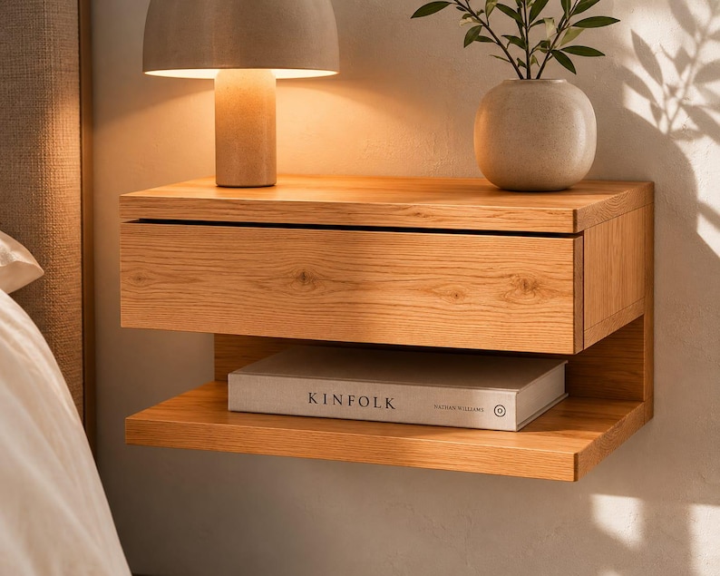 May include: A wall-mounted light oak nightstand featuring a single horizontal drawer and an open lower shelf. A white book with the text KINFOLK is placed on the shelf. The wood grain is visible across the front of the drawer and shelf.