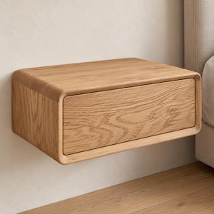 May include: A wall-mounted floating nightstand crafted from light-toned oak wood. The rectangular unit features a single front-facing drawer with a smooth, rounded edge design and a visible natural wood grain pattern throughout the surface.