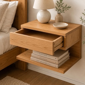 May include: A wall-mounted light oak nightstand featuring a single pull-out drawer and an open lower shelf. The shelf holds three stacked books titled The Kinfolk Home, Kinfolk, and Live Beautifully, with a lamp and bowl on top.