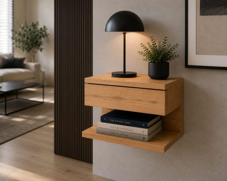May include: A wall-mounted light oak floating nightstand featuring a single drawer and an open lower shelf. A black dome-shaped table lamp and a small potted green plant sit on top, with three stacked books resting on the lower shelf.