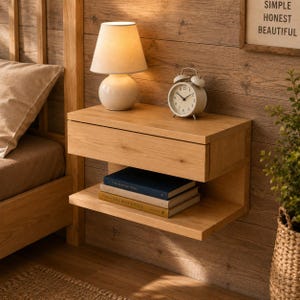 May include: A light oak floating nightstand with a single drawer and an open lower shelf. On top sits a white table lamp and a silver analog alarm clock. Two books are stacked on the lower shelf. The unit is mounted on a wood-paneled wall.