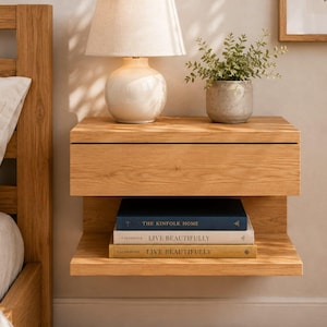 May include: A wall-mounted light oak nightstand featuring a single top drawer and an open lower shelf. The shelf holds three stacked books titled The Kinfolk Home and Live Beautifully. The wood grain is visible throughout the rectangular structure.