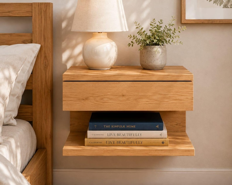 May include: A wall-mounted light oak nightstand featuring a single top drawer and an open lower shelf. The shelf holds three stacked books titled The Kinfolk Home and Live Beautifully. The wood grain is visible throughout the rectangular structure.