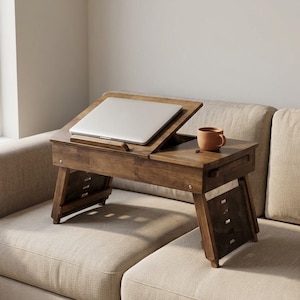 Op de afbeelding: Een houten laptoptafel met een kantelbaar oppervlak en een kleine lade. Een zilveren laptop ligt op de tafel en een bruine mok staat ernaast. De tafel staat op een beige bank.