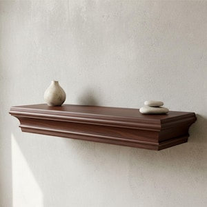 Può includere: Una mensola galleggiante in legno marrone scuro con un bordo decorativo è montata su una parete di colore chiaro. Un piccolo vaso bianco sporco e due pietre grigie lisce poggiano sulla mensola, aggiungendo un tocco di arredamento minimalista.