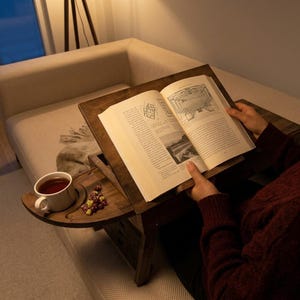 Op de afbeelding: Een houten boekenstandaard met een ingebouwde bijzettafel. Een open boek ligt op de standaard, met een kopje thee en snoepjes op de bijzettafel. De standaard staat op een beige bank.