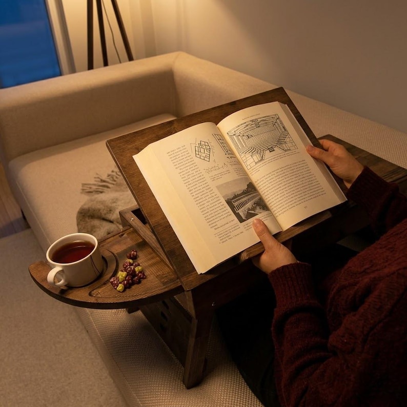 Op de afbeelding: Een houten boekenstandaard met een ingebouwde bijzettafel. Een open boek ligt op de standaard, met een kopje thee en snoepjes op de bijzettafel. De standaard staat op een beige bank.
