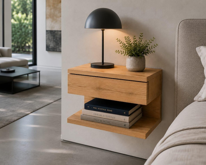 May include: A wall-mounted light oak nightstand featuring a single drawer and an open lower shelf. A black dome-shaped table lamp and a small potted green plant sit on top, while three stacked books rest on the lower shelf.