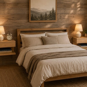 May include: A wooden bed frame with a light beige duvet and a brown throw blanket. Two floating wooden nightstands hold white lamps and a clock. A framed landscape print hangs on the wood-paneled wall above the headboard.