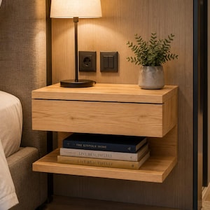 May include: A wall-mounted light wood floating nightstand with a single drawer and an open lower shelf. The shelf holds three stacked books titled The Kinfolk Home and Live Beautifully. A black lamp and a small potted plant sit on top.