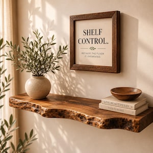 May include: A live-edge wooden floating shelf holding a speckled ceramic vase with greenery, a small wooden bowl, and two stacked books. Above the shelf hangs a square wooden frame containing the text: SHELF CONTROL. BECAUSE THE FLOOR IS OVERRATED.