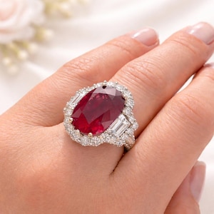 May include: A large, oval-cut ruby ring with a halo of small diamonds and baguette-cut diamonds on the sides. The ruby is a deep red color, and the ring is set in a gold band. The ring is on a person's finger.