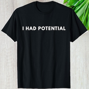 May include: Black crew neck t-shirt with the white text "I HAD POTENTIAL" across the chest. The shirt is made of a soft material and has short sleeves. The background includes green leaves.