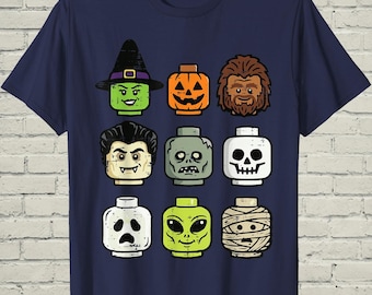 May include: A navy blue t-shirt featuring a graphic print of nine stylized toy-style heads arranged in a grid. The characters include a witch, a jack-o'-lantern, a hairy creature, a vampire, a zombie, a skull, a ghost, an alien, and a mummy.