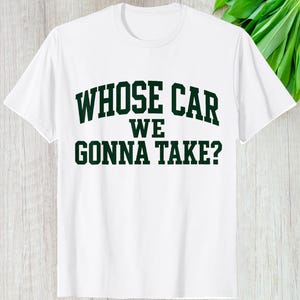 May include: White t-shirt with the text "WHOSE CAR WE GONNA TAKE?" in dark green, block letters. The shirt has a crew neck and short sleeves. The text is centered on the front of the shirt.