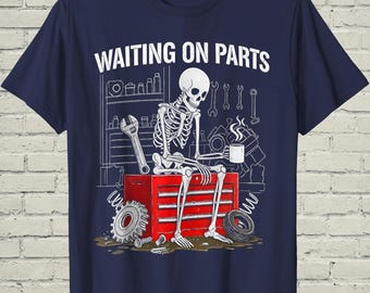 May include: A navy blue t-shirt featuring a white graphic of a skeleton sitting on a red tool chest, holding a steaming mug. The text WAITING ON PARTS is printed above. The background shows a garage shelf with tools, a gear, and a tire.