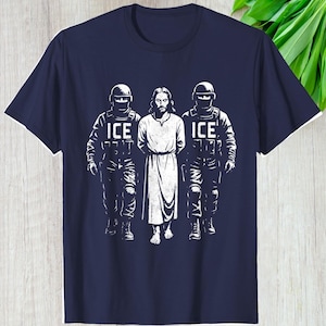 May include: Navy blue t-shirt featuring a graphic of Jesus flanked by two figures in tactical gear, each with the word "ICE" printed on their uniforms. The graphic is in white, creating a stark contrast against the dark blue fabric.