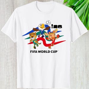 May include: White t-shirt featuring the FIFA World Cup logo and three cartoon mascots: an eagle, a jaguar, and a deer-like creature. The mascots are positioned above the text "FIFA WORLD CUP". The shirt has a crew neck and short sleeves.