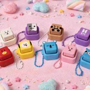 May include: A collection of nine square-shaped character keychains in various colors including red, yellow, blue, pink, purple, white, and brown. Each keychain features a unique cartoon face or the BTS logo, with a small ball chain attached.