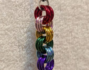 Puede incluir: Un collar colorido con una cadena de anillos entrelazados en colores del arcoíris: rojo, rosa, dorado, verde, azul y morado. La cadena está suspendida de una cadena de metal negro.